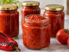 Четири буркана с домашна лютеница без патладжан, поднесени до печени червени чушки и пресни домати.