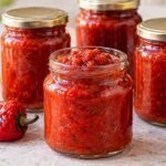 Четири буркана с домашна лютеница с мед и печени чушки, подредени до печени червени чушки, домати и бурканче с мед.